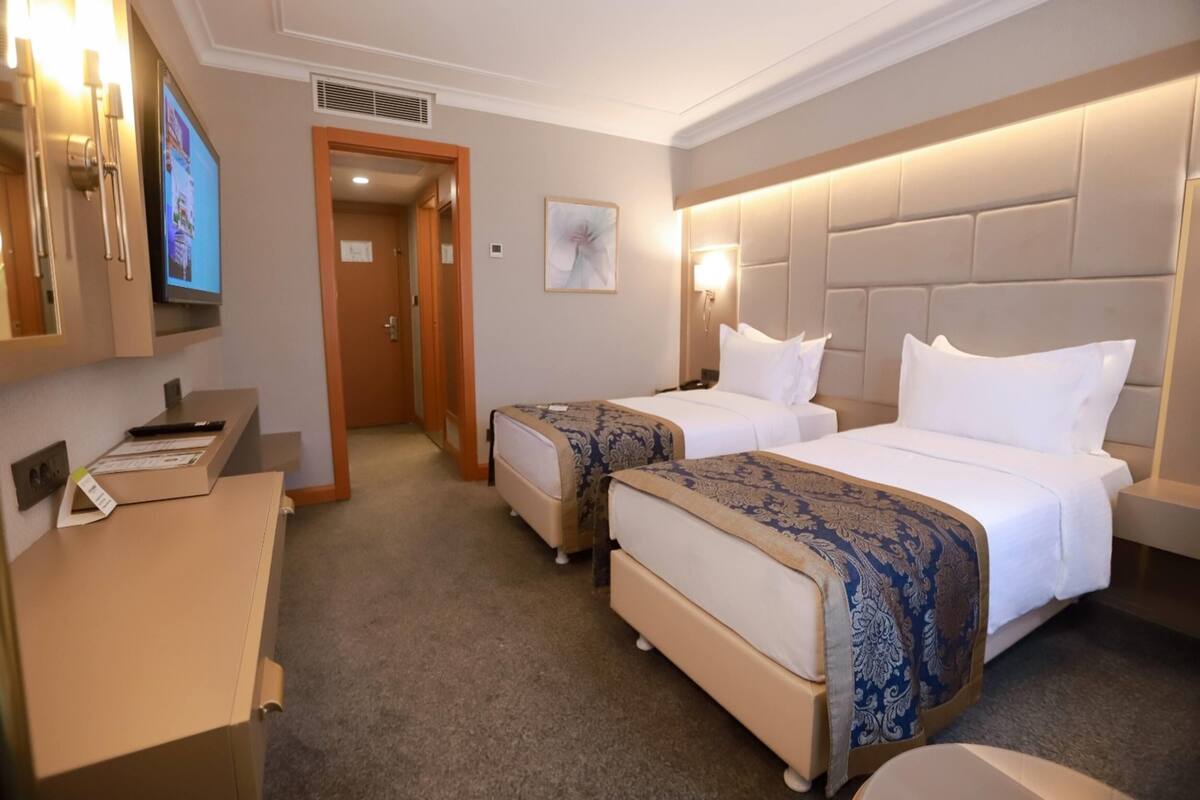 Ein Hotelzimmer mit zwei Betten, einem Schreibtisch mit Computer, einem Fernseher und einer Tür zu einem anderen Raum.
