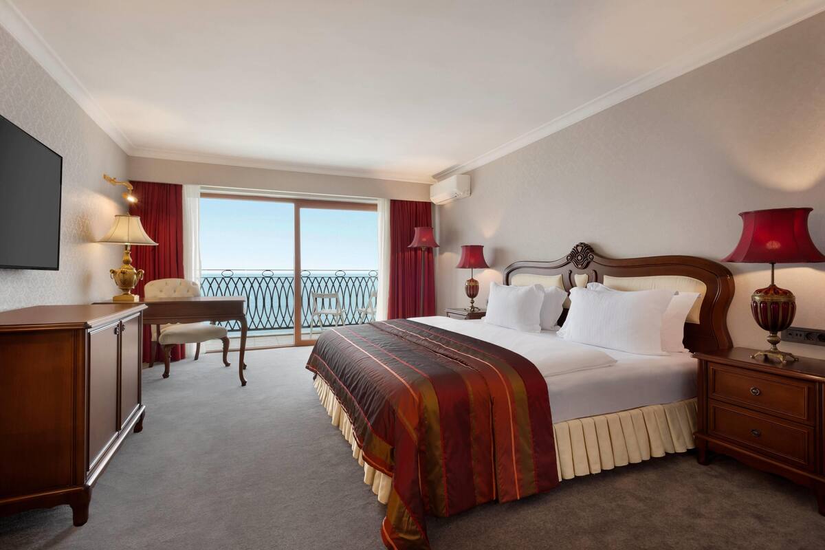 Ein Hotelzimmer mit einem großen Bett, einem Schreibtisch, einem Stuhl und einem Balkon mit roten Vorhängen.