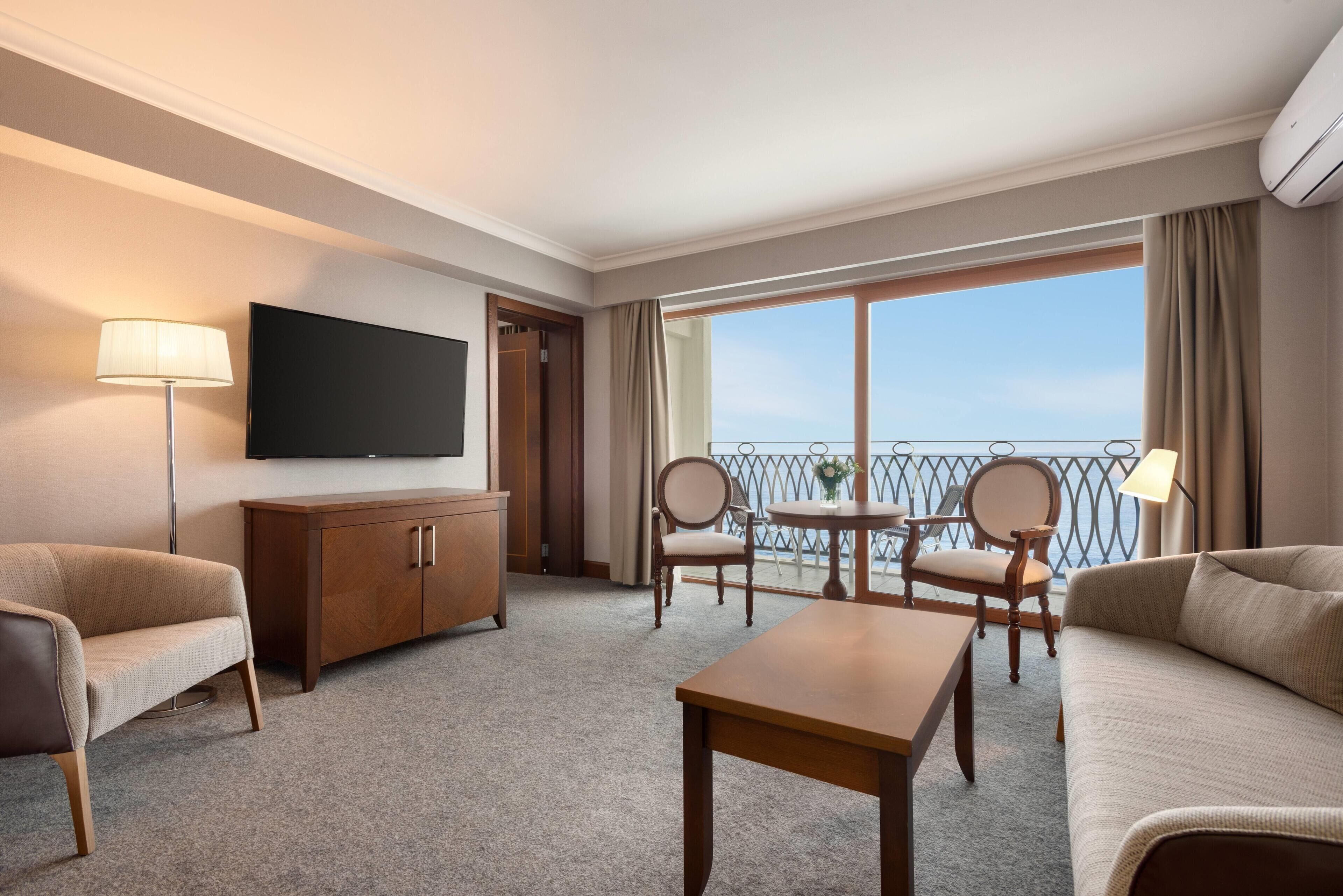 A hotel room with a flat-screen TV, a sofa, a chair, a small table, and a balcony with a railing.