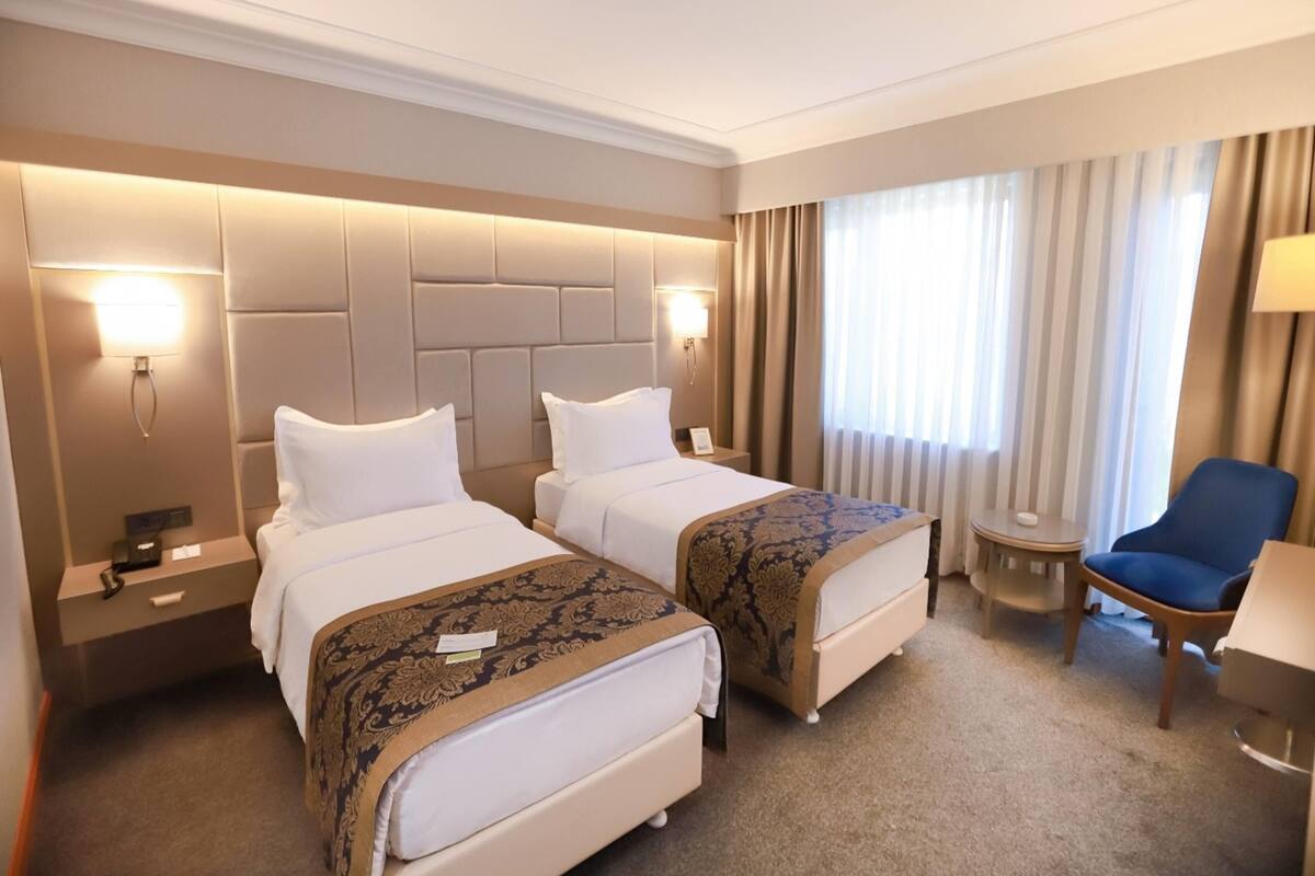 Ein Hotelzimmer mit zwei Betten, einem kleinen Tisch, einem Sessel und einem großen Fenster mit Vorhängen.