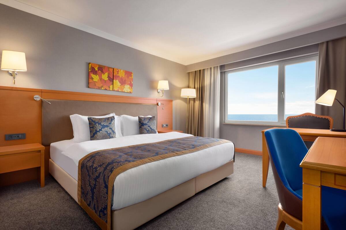 Ein Hotelzimmer mit einem großen Bett, zwei Nachttischlampen, einem Schreibtisch mit Stuhl, einem Fenster mit Vorhängen und einem farbenfrohen Bild an der Wand.