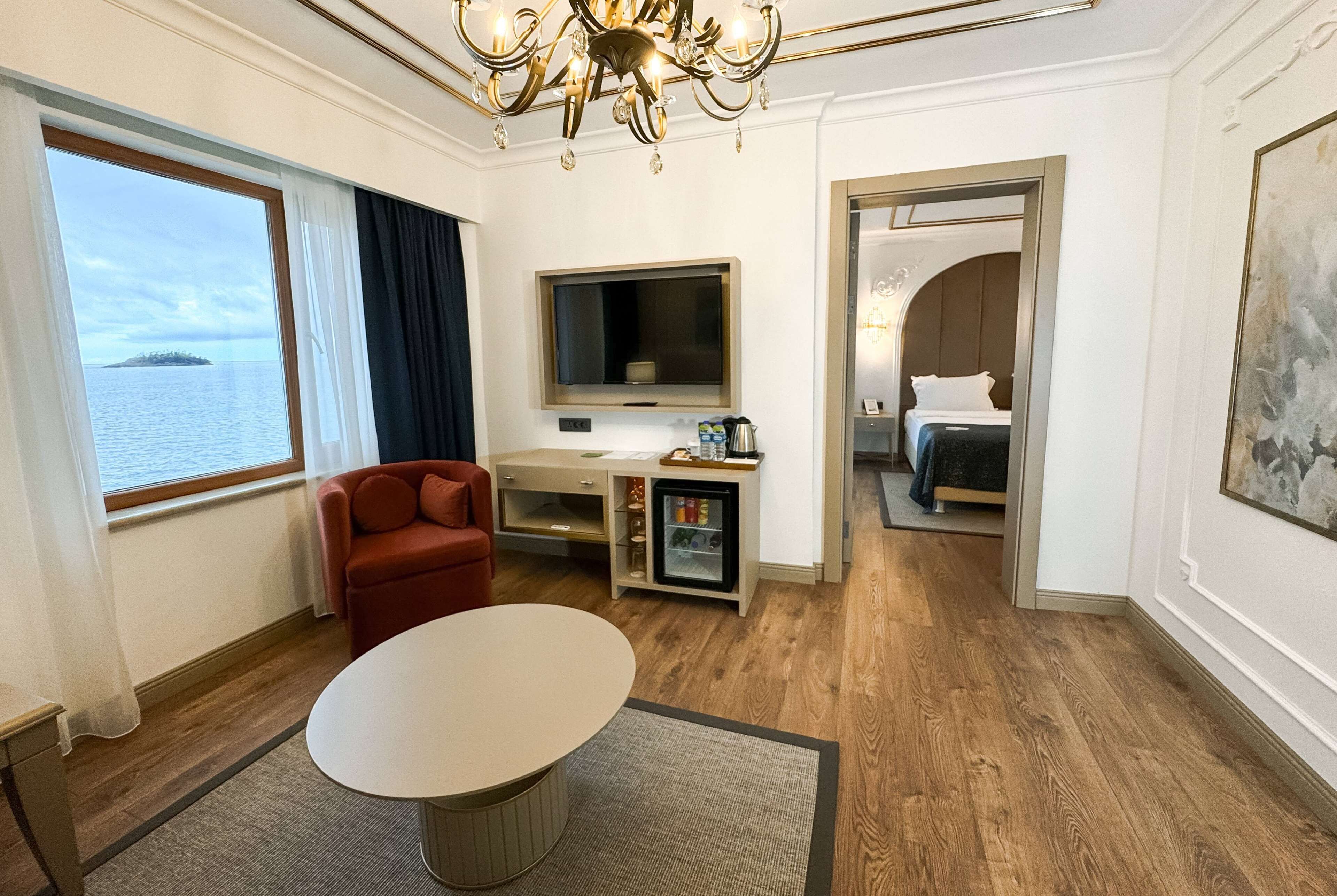 Ett hotellrum med ett stort fönster, en TV, ett litet kylskåp, en röd fåtölj, ett runt bord och en säng med en sänggavel.