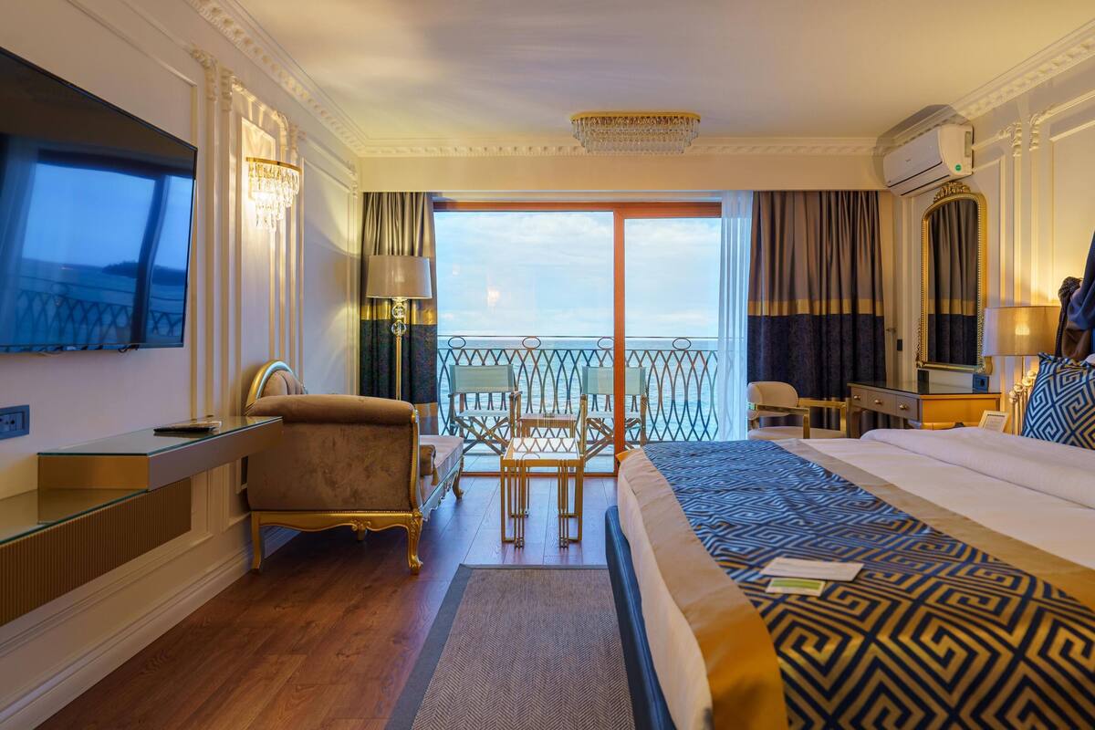 Ein Hotelzimmer mit Bett, Schreibtisch, Sessel, Fernseher und einem Balkon mit Aussicht, sowie einer Klimaanlage.