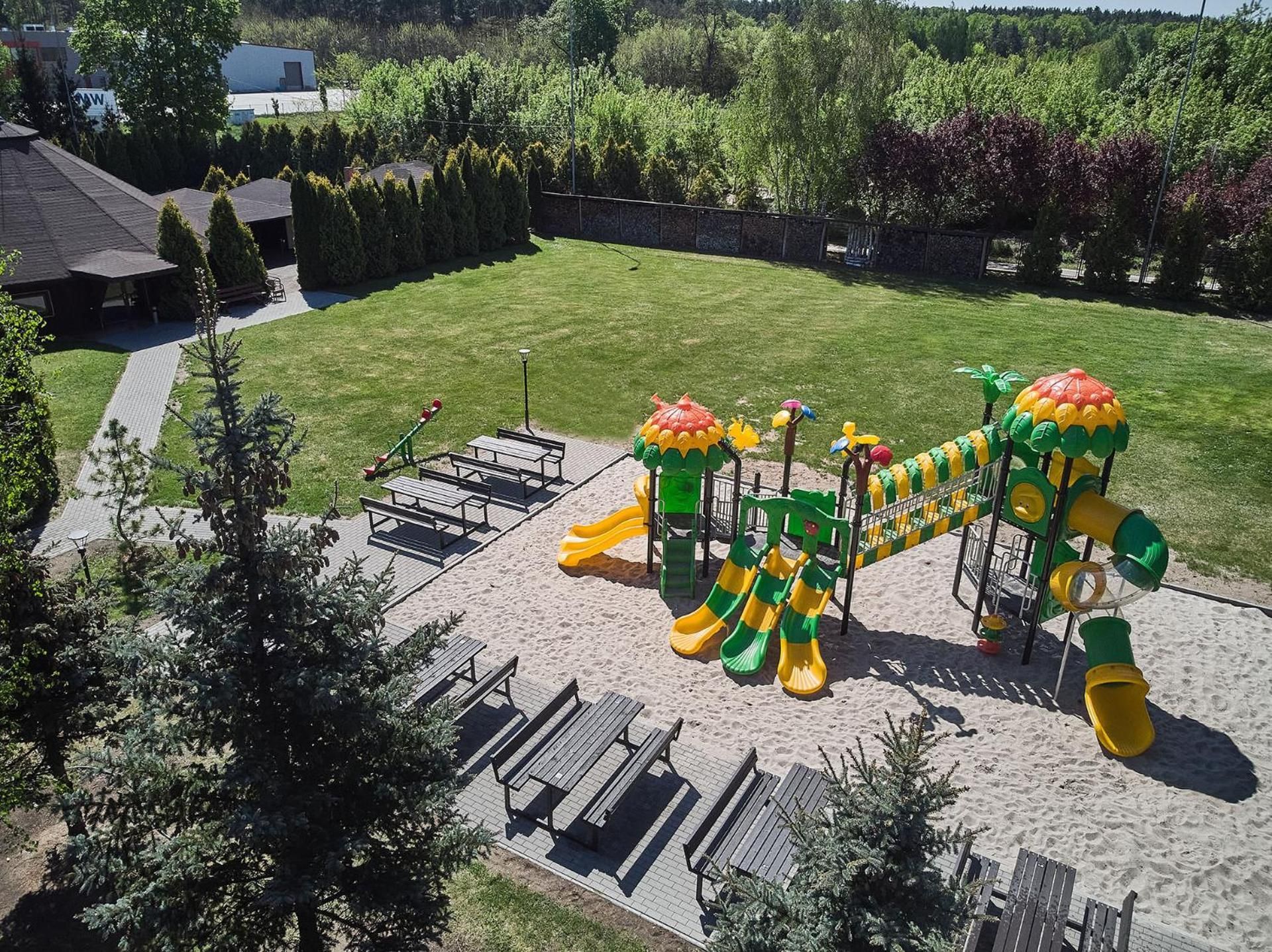 Une aire de jeux avec des toboggans et des balançoires, entourée d’une zone pavée et d’arbres.