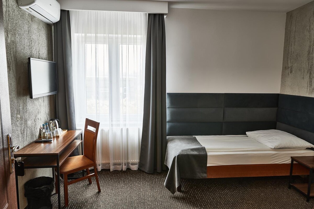 A hotel room with a bed, a desk, a chair, a TV, and a window with curtains.