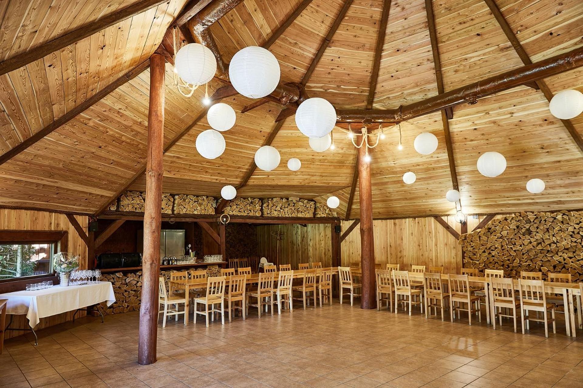 A rustic wooden hall with a high ceiling, multiple tables and chairs, and hanging spherical light fixtures.