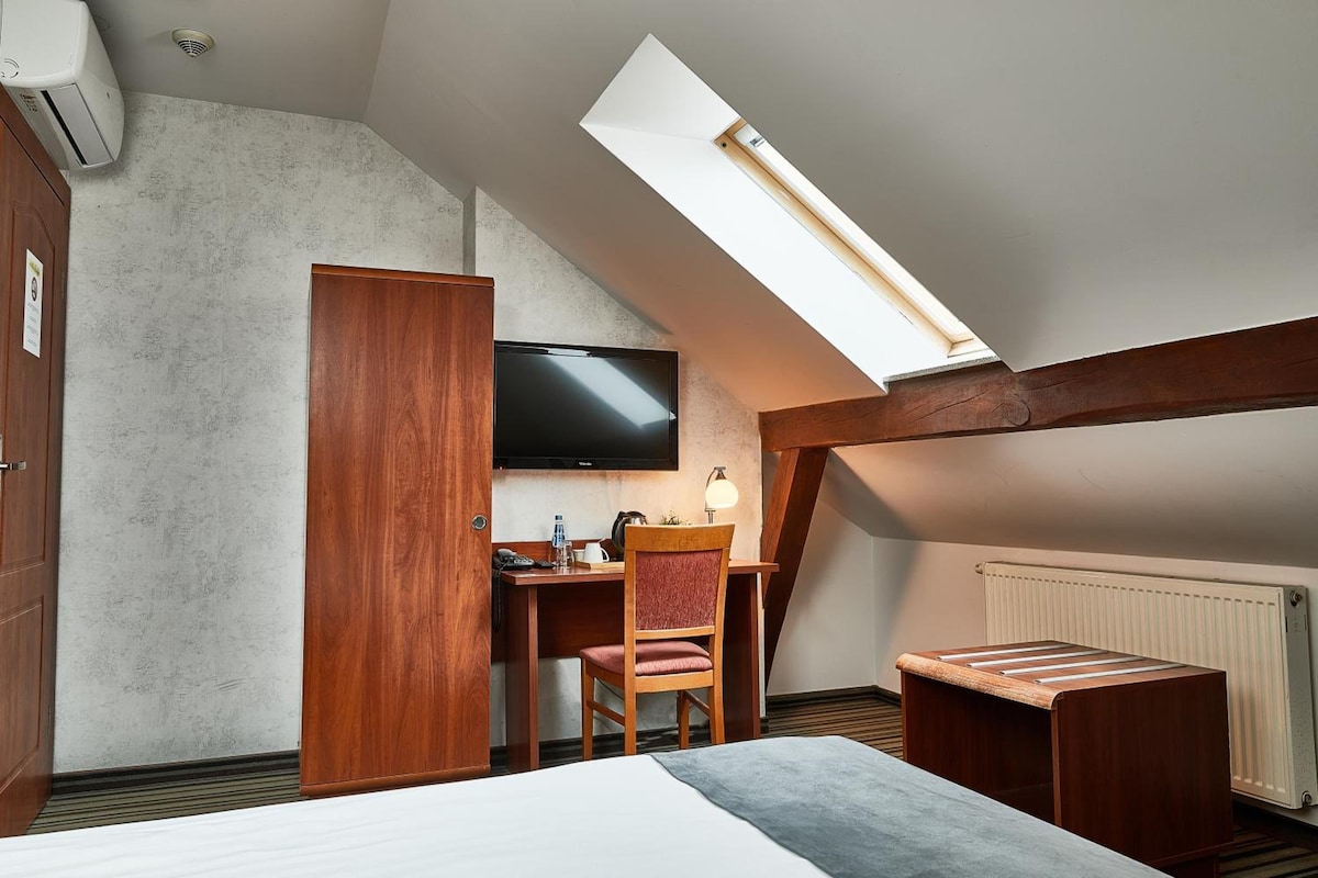 Una habitación con techo inclinado, una cama, un escritorio con silla, un televisor y un radiador.
