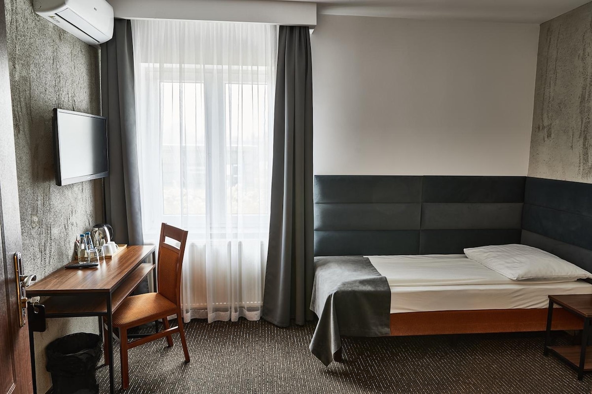 Una habitación de hotel con cama, escritorio, silla, televisión y ventana con cortinas.