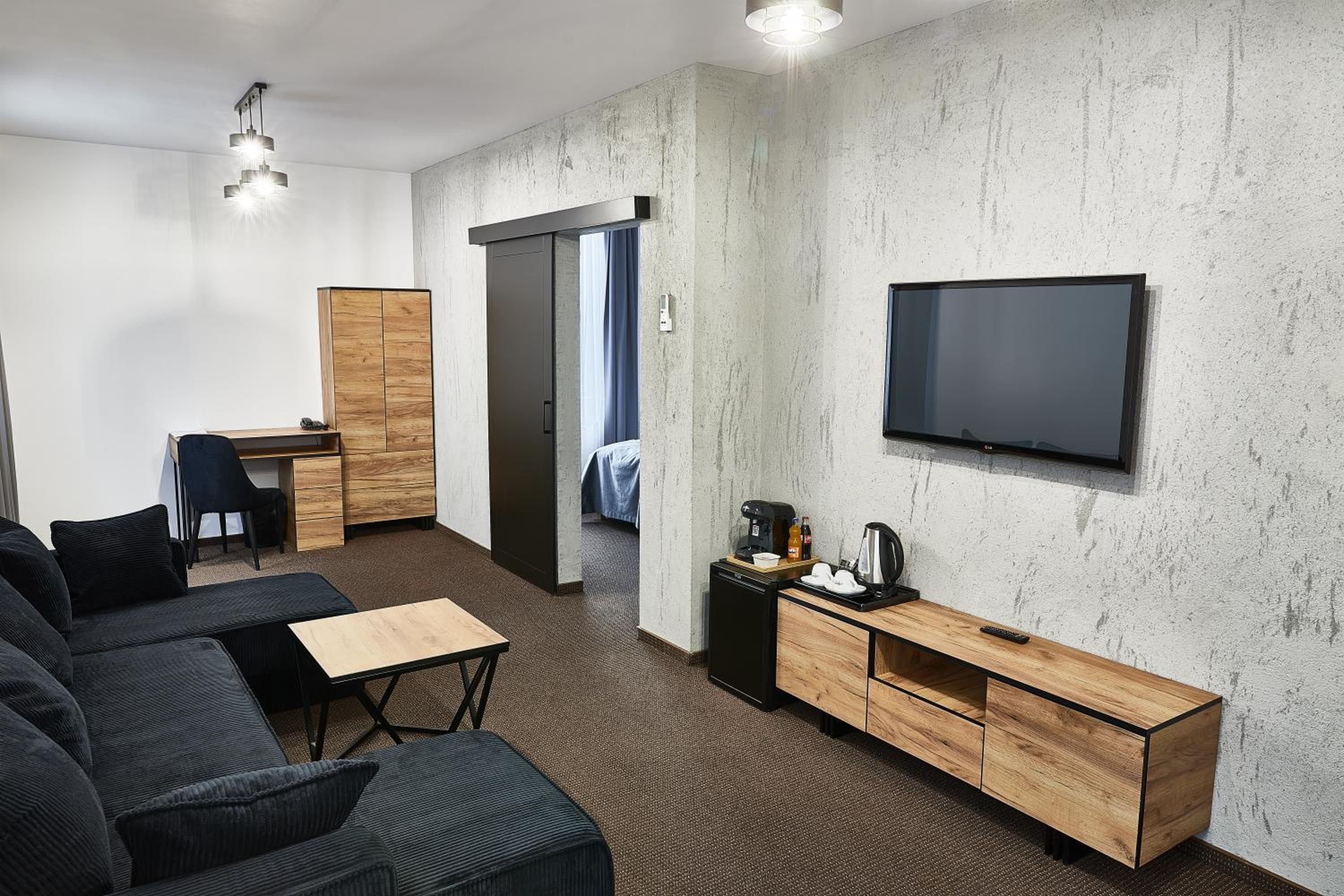 A modern hotel room with a sofa, a desk, a television, and a kitchenette.