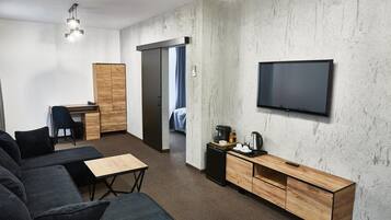 A modern hotel room with a sofa, a desk, a television, and a kitchenette.