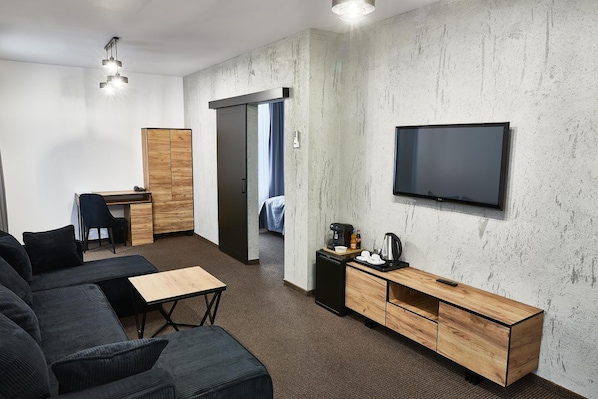 A modern hotel room with a sofa, a desk, a television, and a kitchenette.