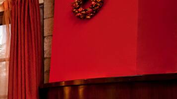 A fireplace with a red mantel and a wreath, surrounded by a wooden floor and red curtains.