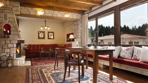 A cozy living room with a stone fireplace, a wooden dining table, and a sofa with red cushions. - Fretzato Traditional Guest House (Pyli)
