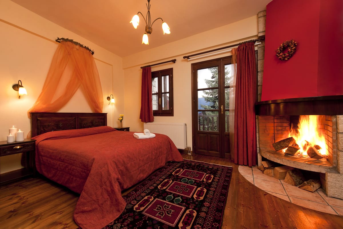 A bedroom with a large bed, a fireplace, a window with curtains, and a nightstand with a lamp.