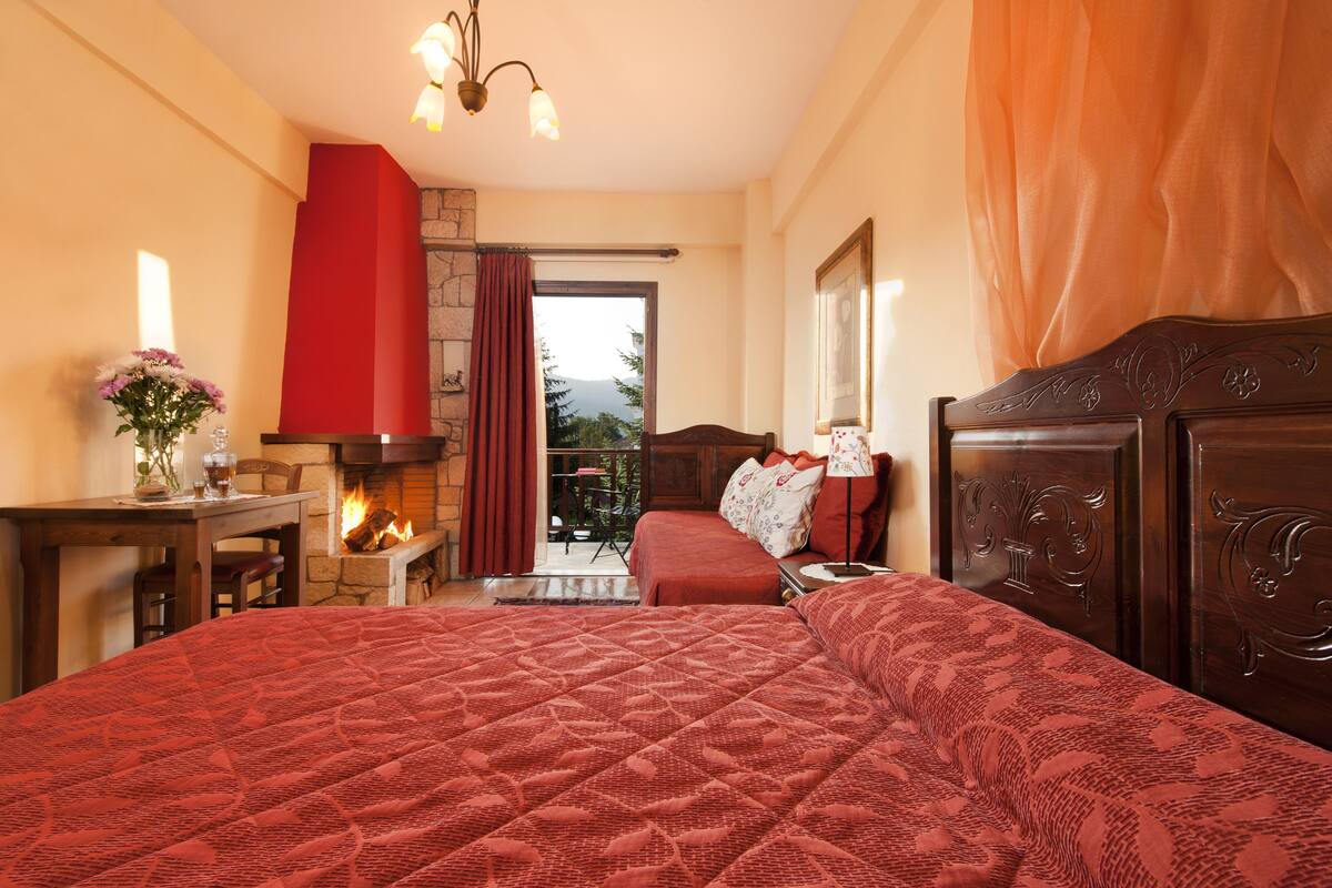 A bedroom with a large bed, a fireplace, a table with a vase of flowers, and a view of the outdoors.