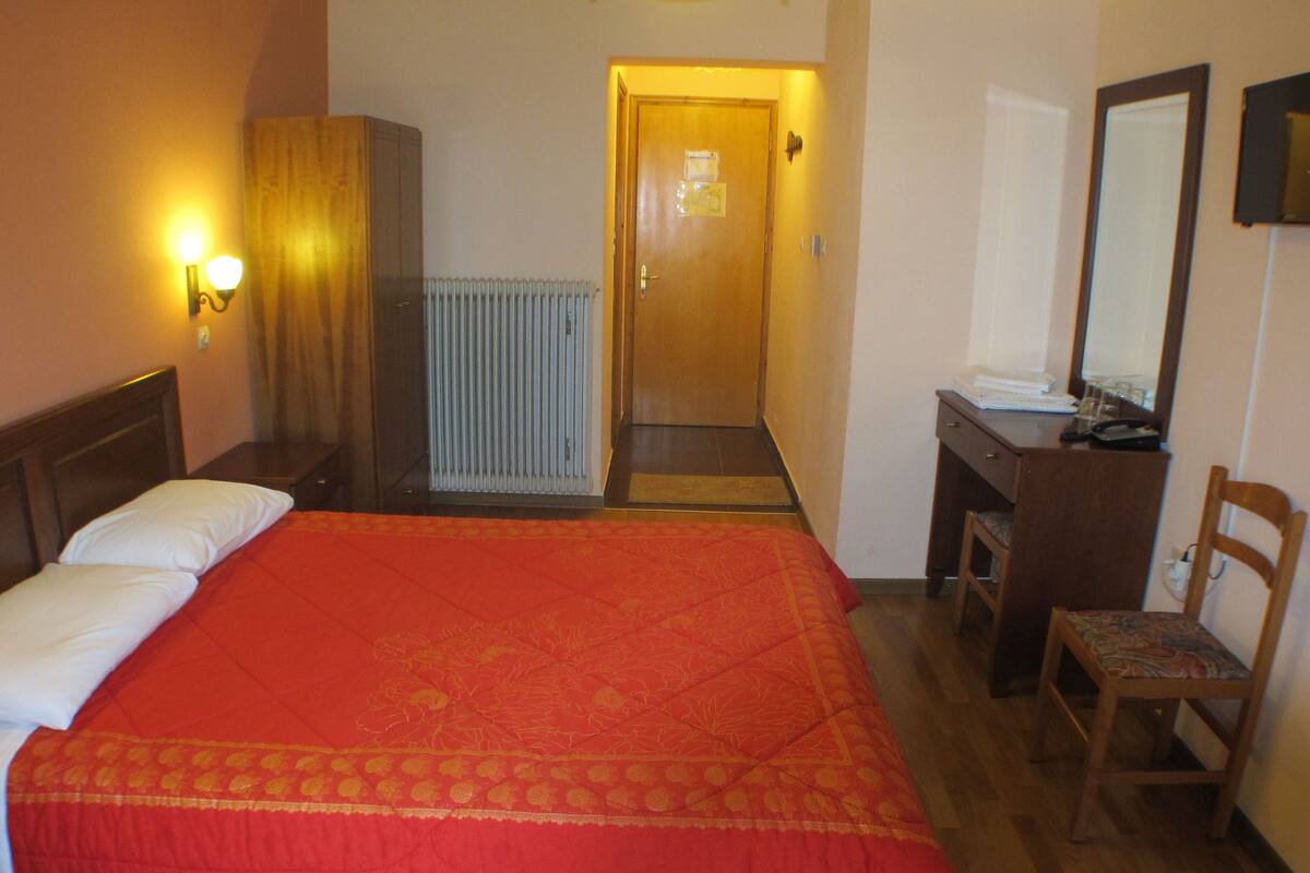 Une chambre d’hôtel avec un grand lit, une armoire en bois, un bureau avec une chaise, un radiateur et une porte donnant sur une autre pièce.