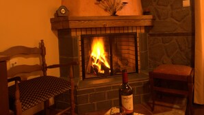 A cozy room with a fireplace, a wooden chair, a table with a bottle and glasses, and a bowl of fruit. - Kroupi Hotel (Pyli)