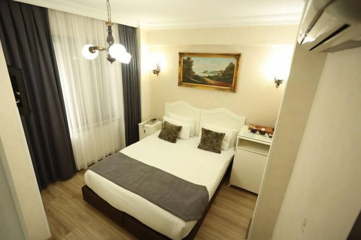 A hotel room with a bed, bedside tables, a painting, and a window with curtains.