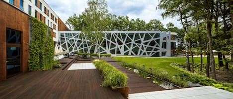 Un edificio moderno con fachada geométrica, rodeado de un jardín cuidado y una terraza de madera.