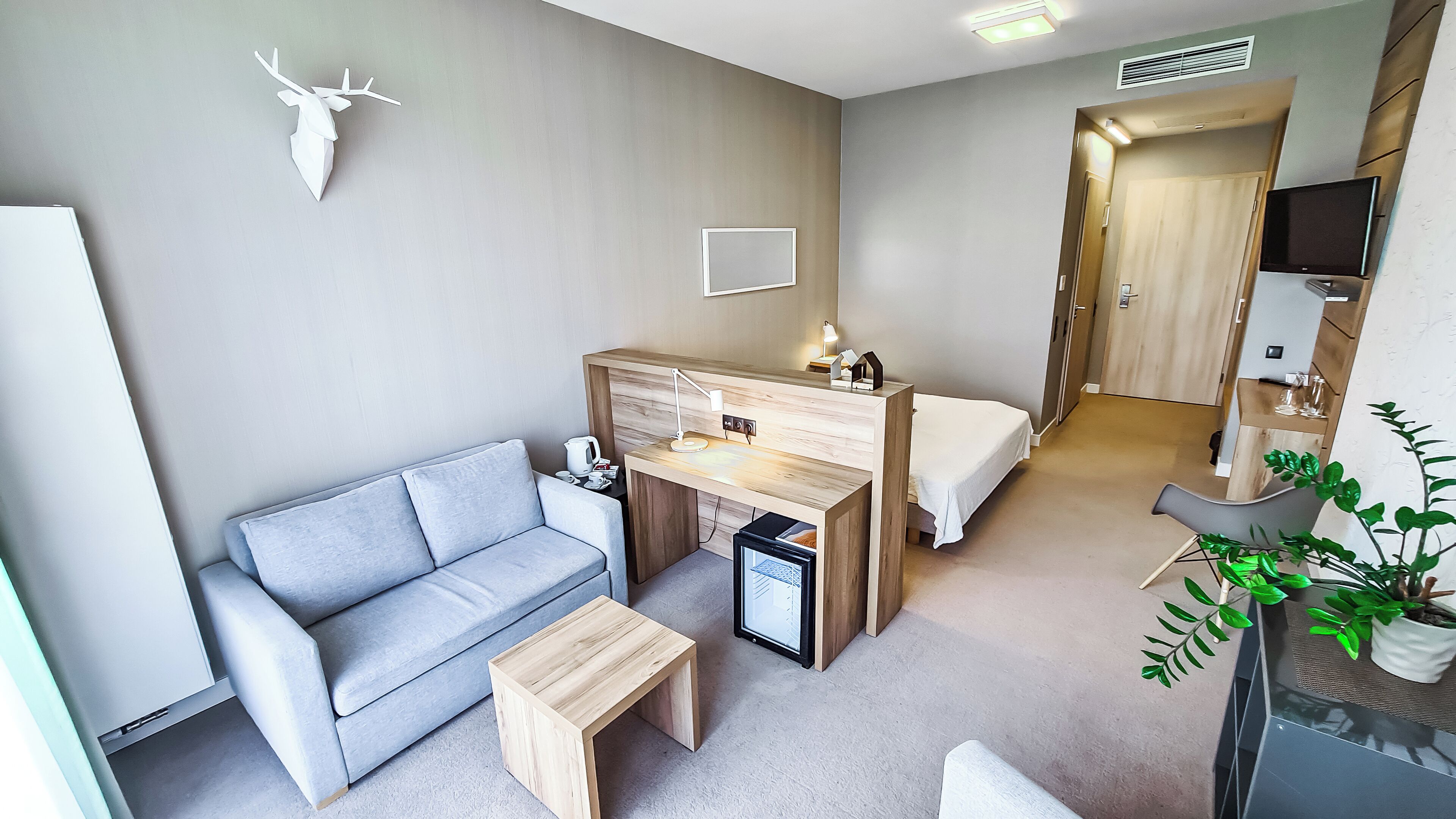 A modern hotel room with a grey sofa, a wooden desk, a small table, a bed, and a TV.