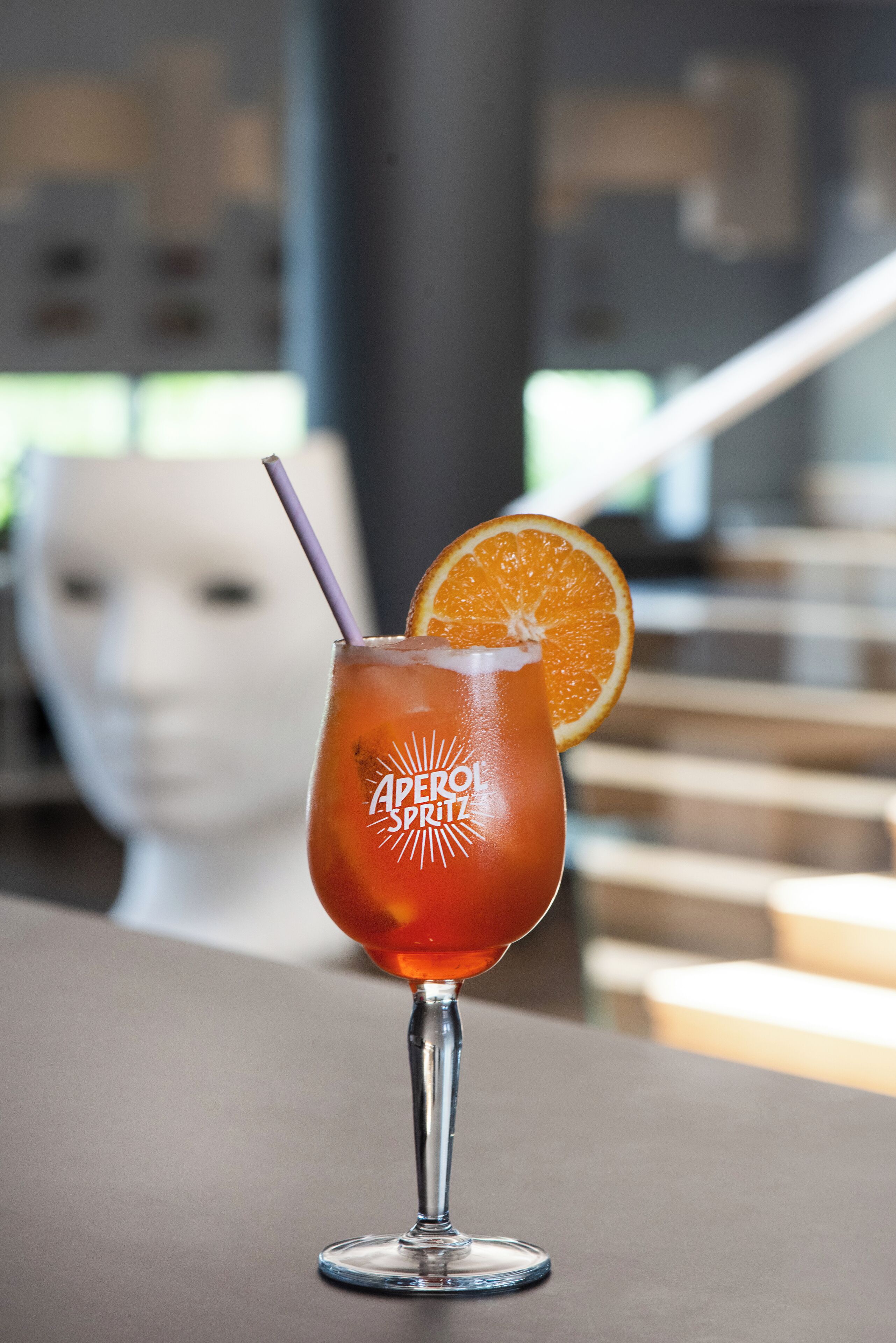Un cóctel con pajita y una rodaja de naranja, etiquetado como "Aperol Spritz", sobre una mesa con un fondo desenfocado.