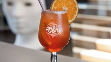 Un cóctel con pajita y una rodaja de naranja, etiquetado como "Aperol Spritz", sobre una mesa con un fondo desenfocado.
