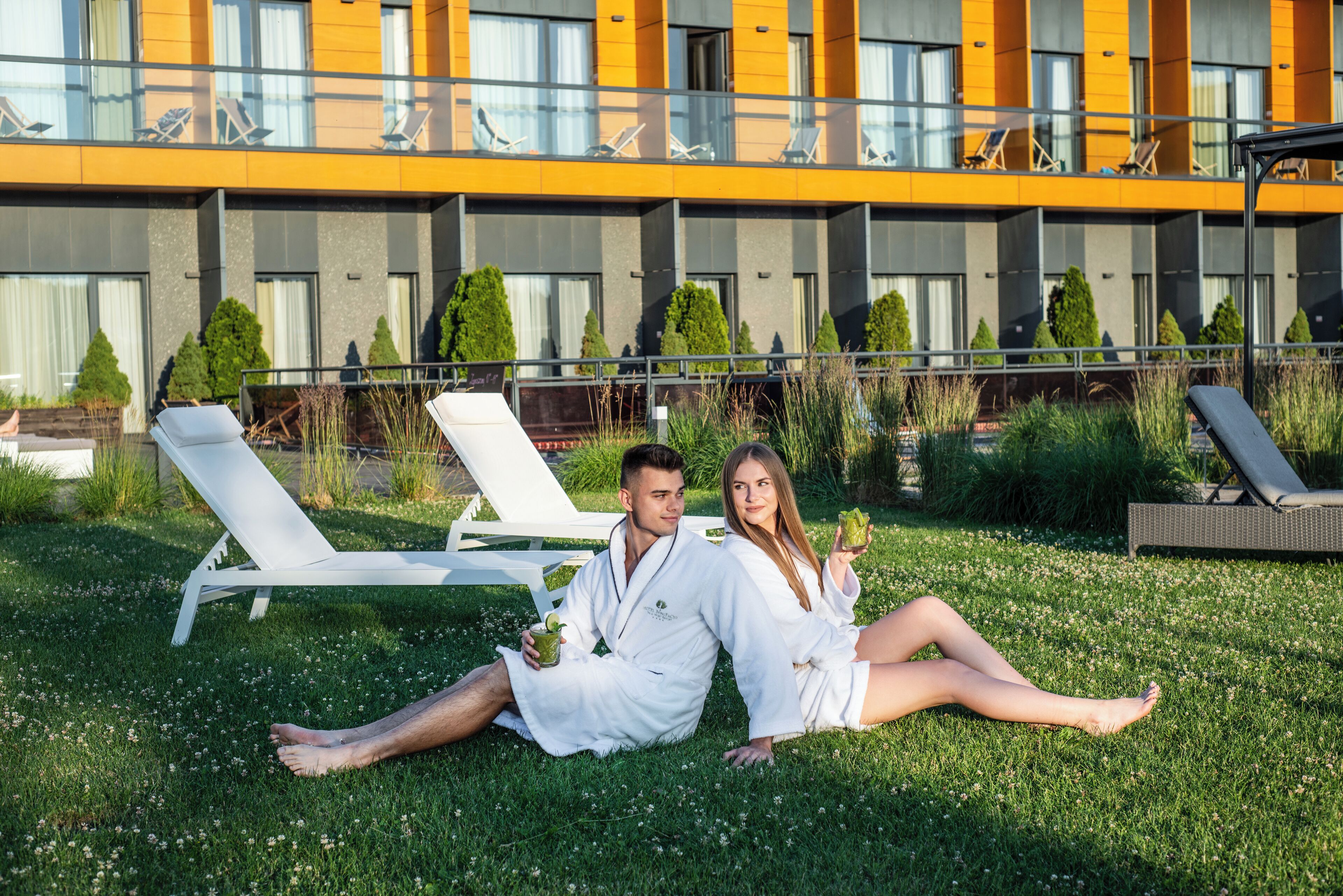 Una pareja descansando sobre el césped frente a un edificio moderno con varios balcones y sillones de descanso.