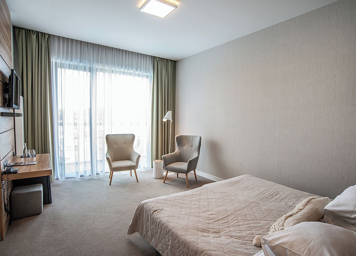 Habitación de hotel con cama, dos sillones, escritorio con computadora y ventana grande con cortinas.