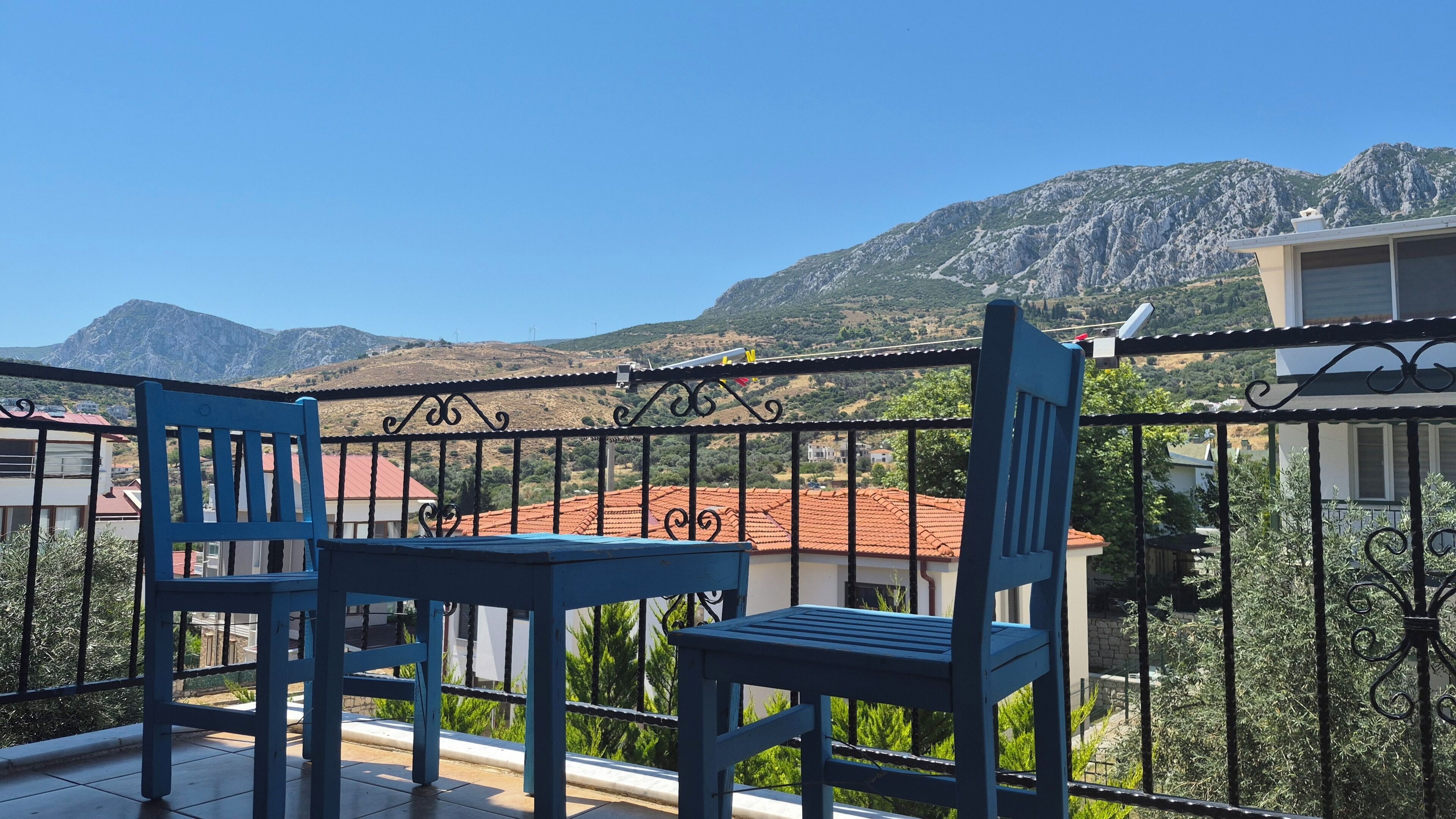 A balcony with two blue chairs and a table, overlooking a mountainous landscape with buildings in the background.