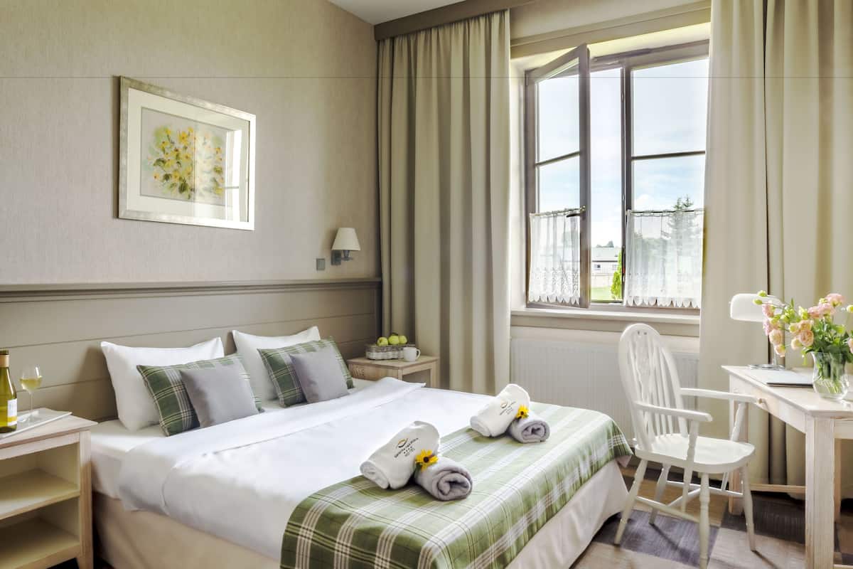 A hotel room with a bed, a desk, a chair, a window with curtains, a nightstand, and a framed picture on the wall.