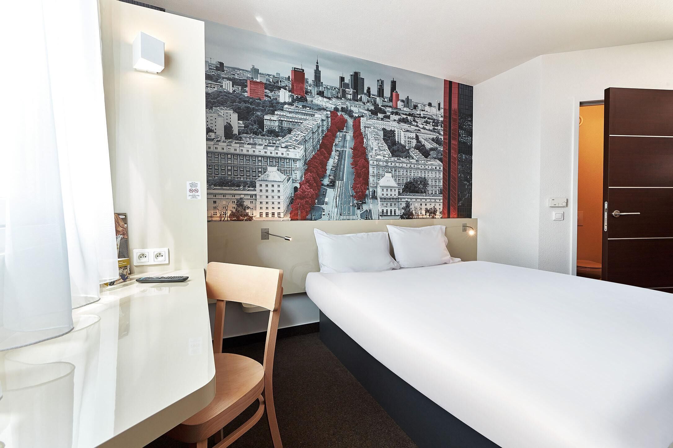 A hotel room with a bed, a desk, a chair, and a cityscape mural on the wall.