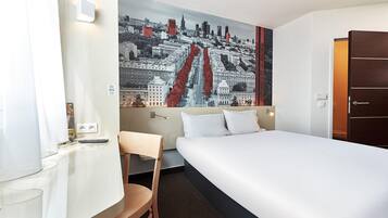 A hotel room with a bed, a desk, a chair, and a cityscape mural on the wall.