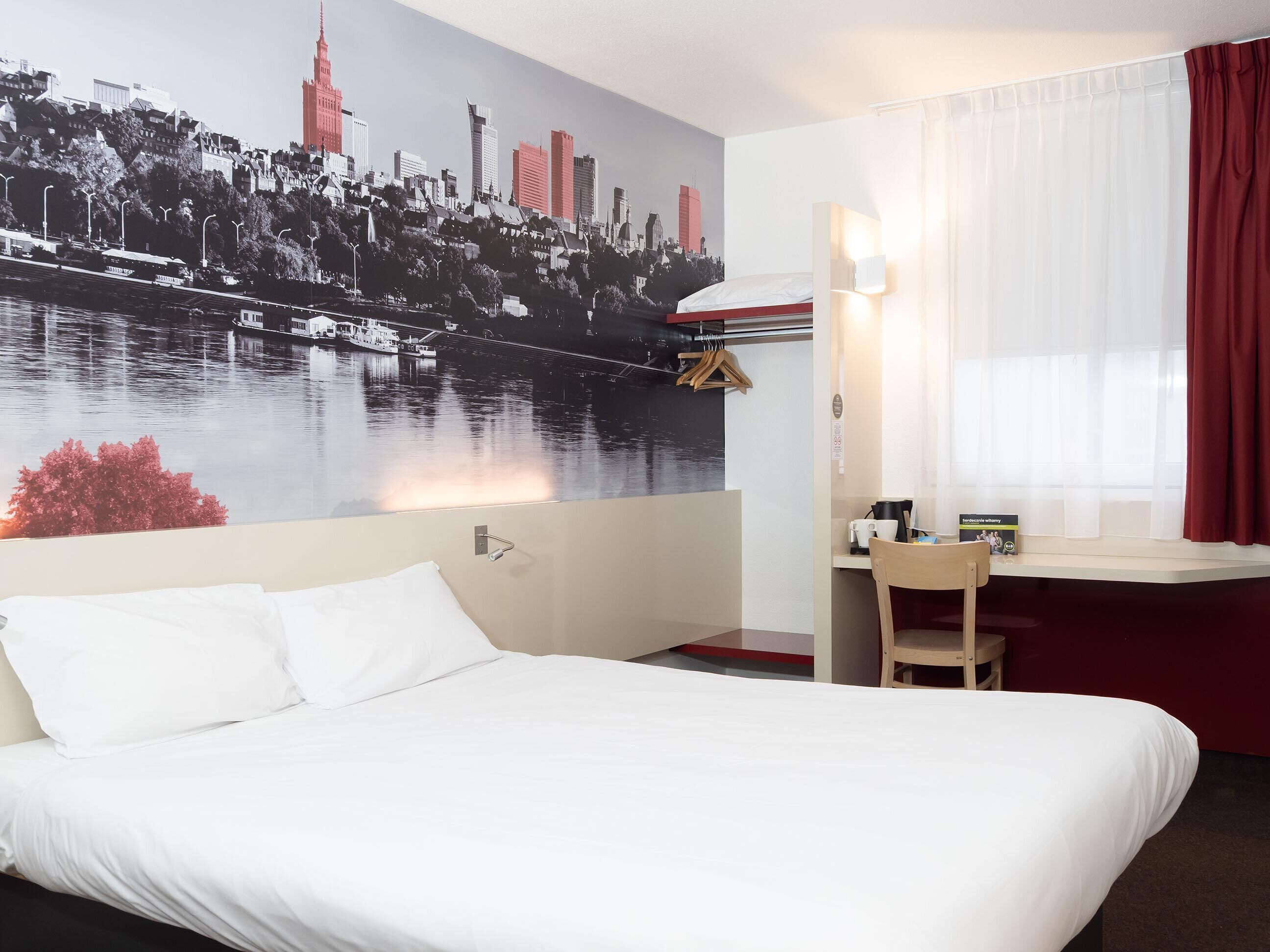 A hotel room with a bed, a desk, a chair, a window with curtains, a wall-mounted light, and a cityscape mural.