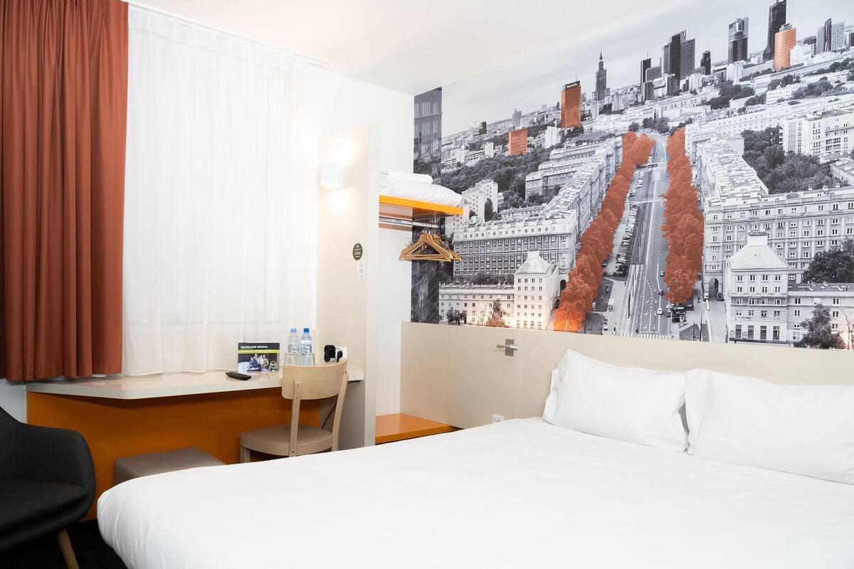 Una habitación de hotel con una cama, un escritorio, una silla, una ventana con cortinas, una lámpara fijada a la pared y un mural con vista a la ciudad.