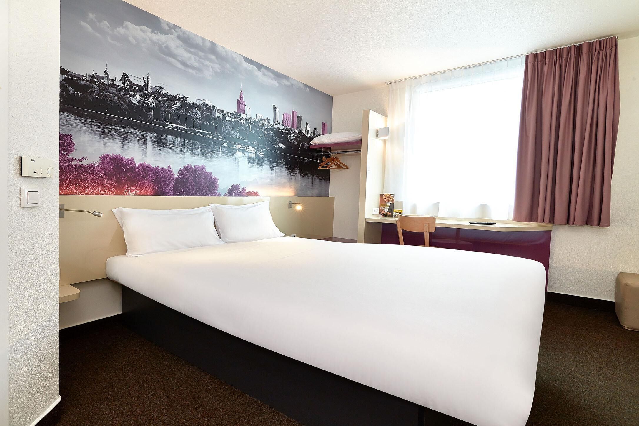 A hotel room with a large bed, a desk with a chair, a window with curtains, and a cityscape mural on the wall.