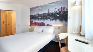 A hotel room with a bed, a desk, a chair, a painting of a cityscape, and a bathroom with a shower and toilet.