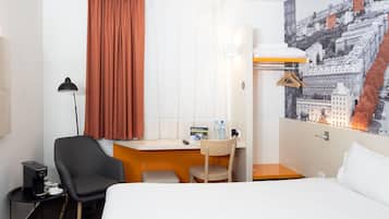 Ett hotellrum med en säng, ett skrivbord med en stol, en lampa, ett litet bord med en kaffebryggare och utsikt över staden.