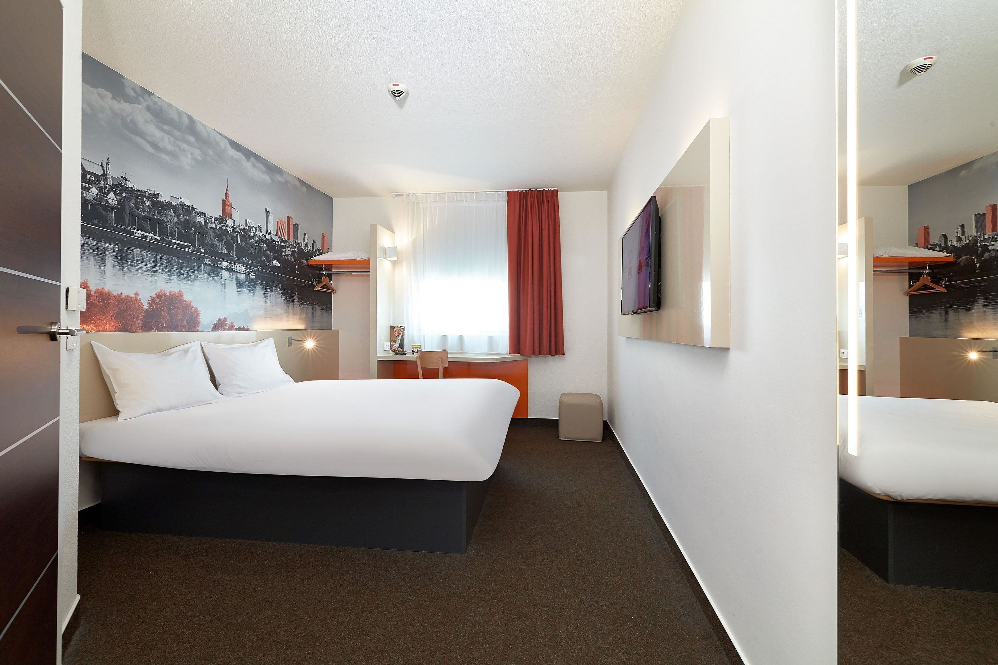 A hotel room with a large bed, a desk, a chair, a TV, and a painting of a cityscape.