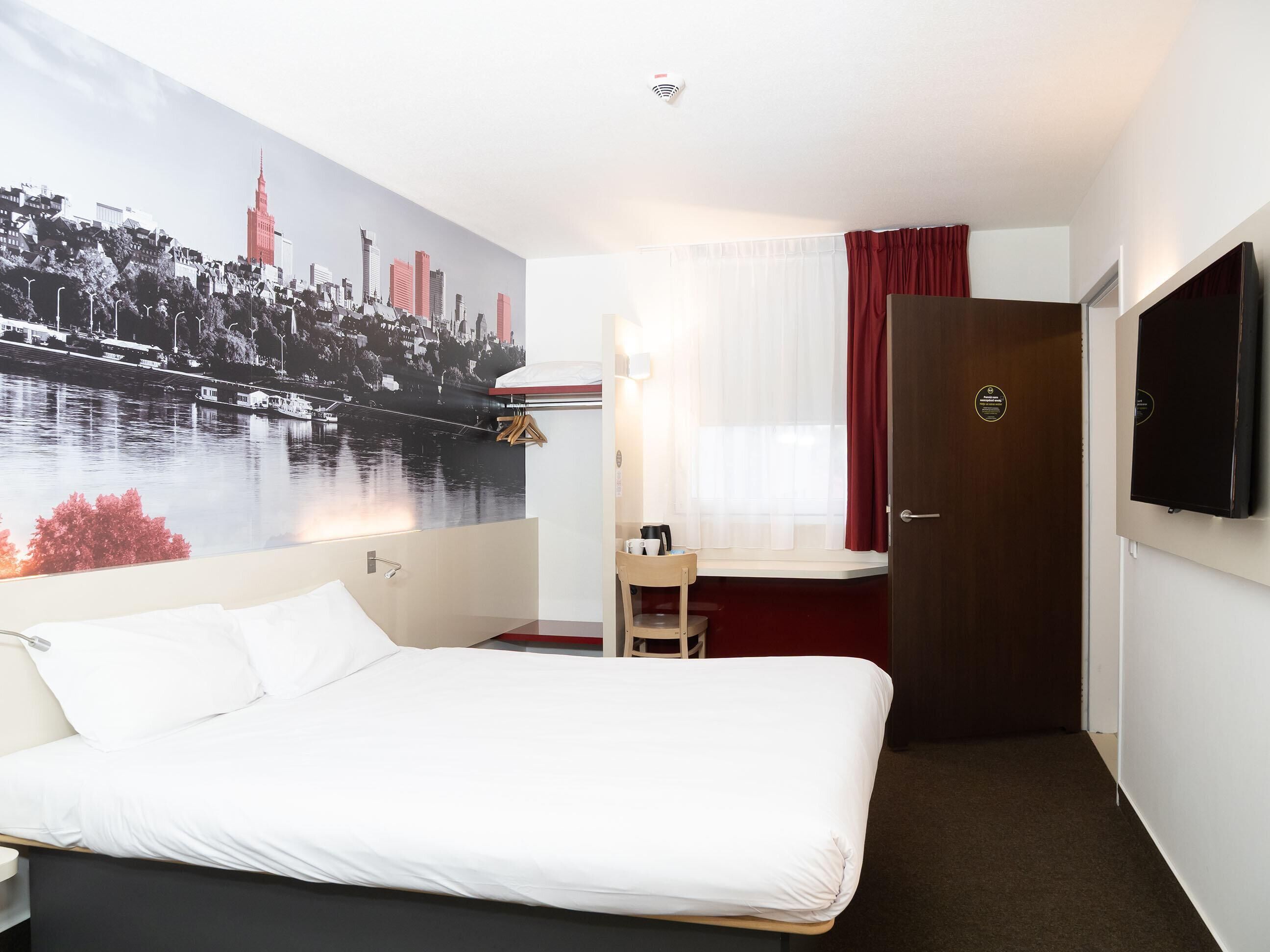 Hotellihuone, jossa on sänky, työpöytä tuolilla, televisio, ikkuna verhoilla ja seinällä kaupunkimaisemaa esittävä seinämaalaus.