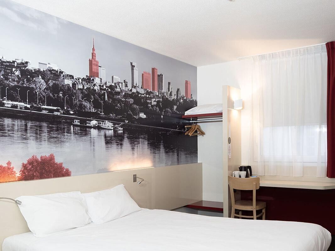 A hotel room with a bed, a desk, a chair, a window with curtains, and a cityscape mural on the wall.