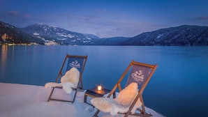 Two lounge chairs with fluffy white cushions on a snow-covered deck overlooking a calm lake with mountains in the background. - Seeglück Hotel Forelle Superior (Millstatt)