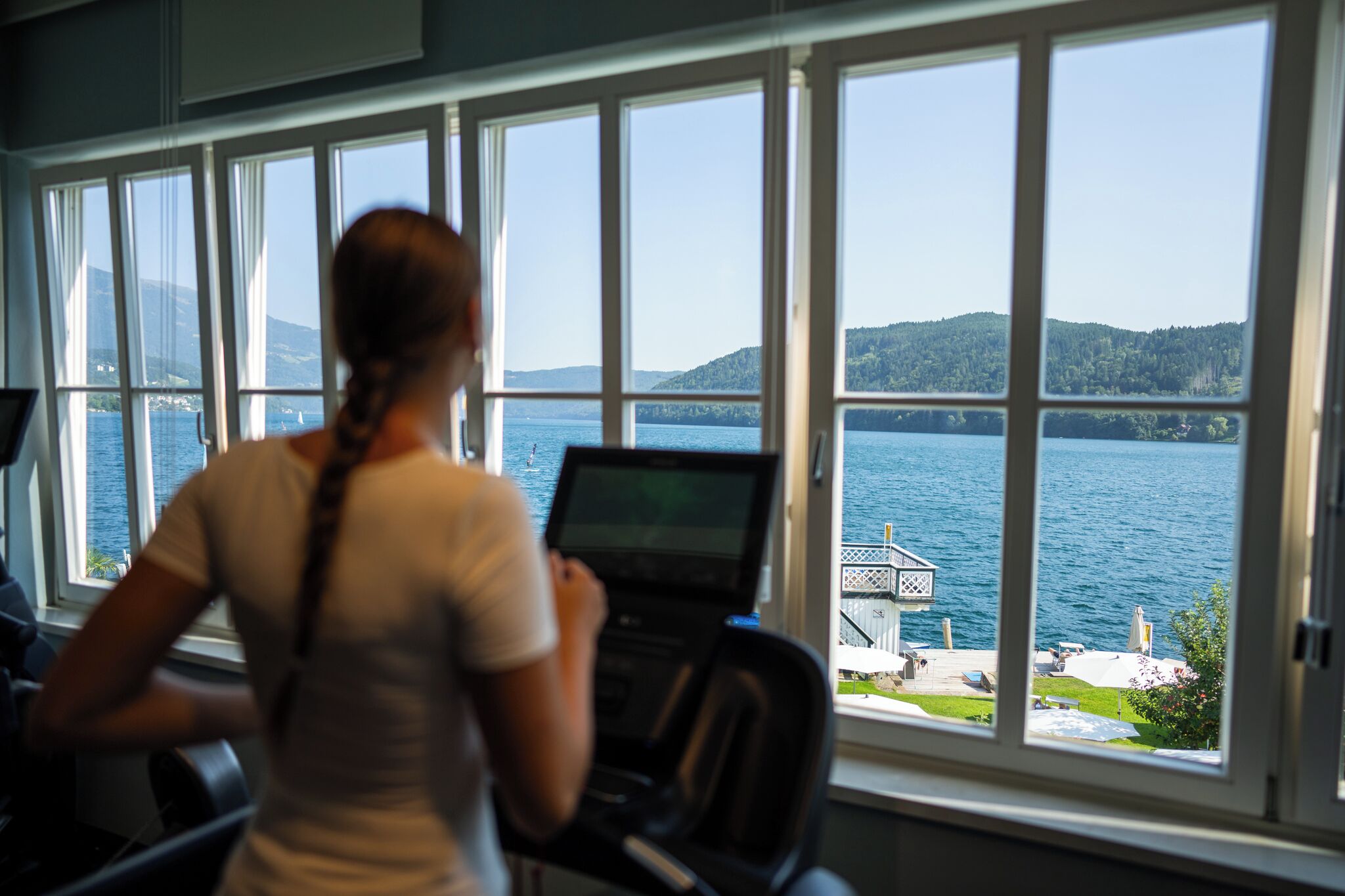 A person using a treadmill with a view of a lake and distant hills through large windows.