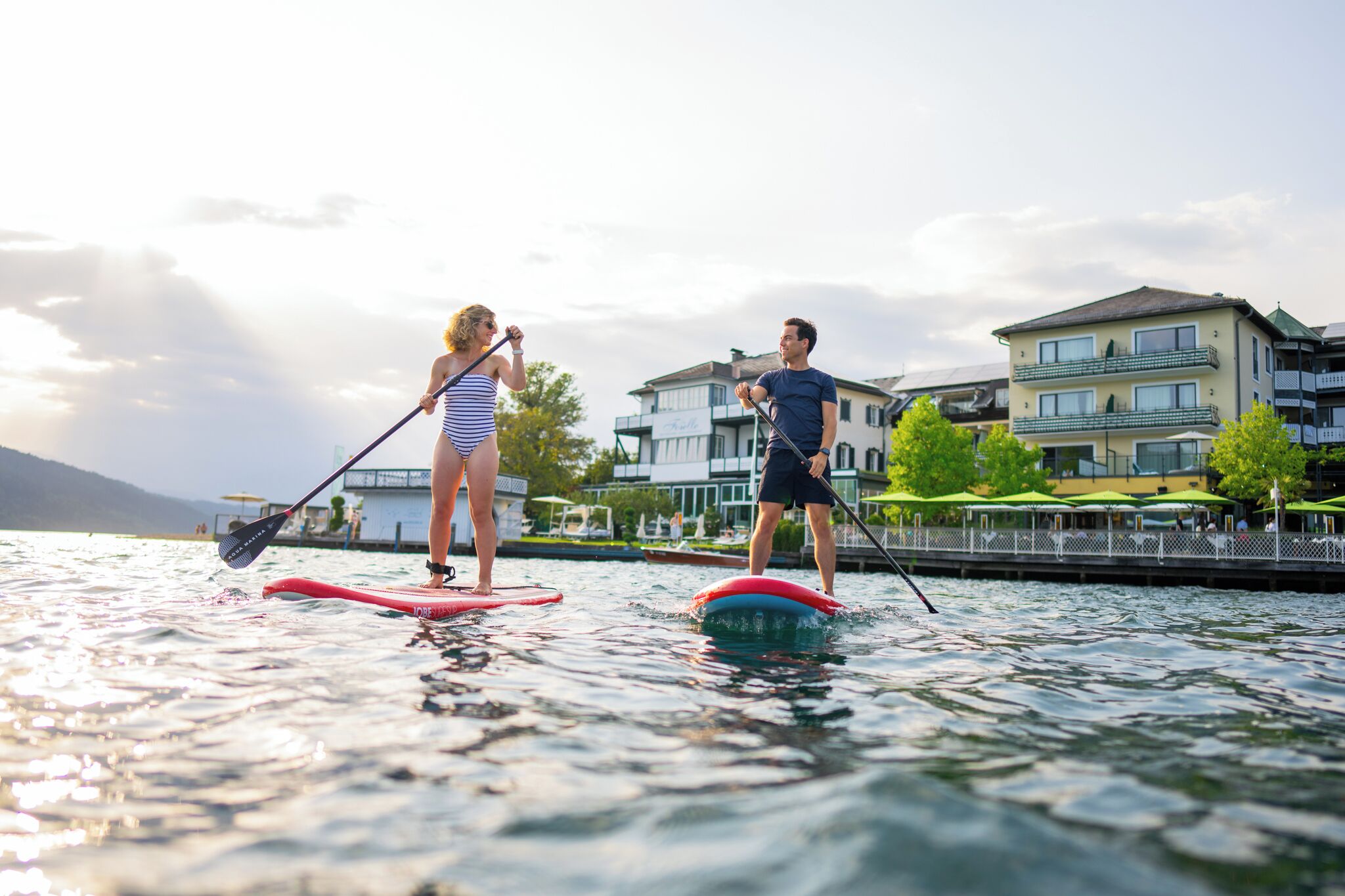 Two people paddleboarding on a calm lake with buildings in the background.
