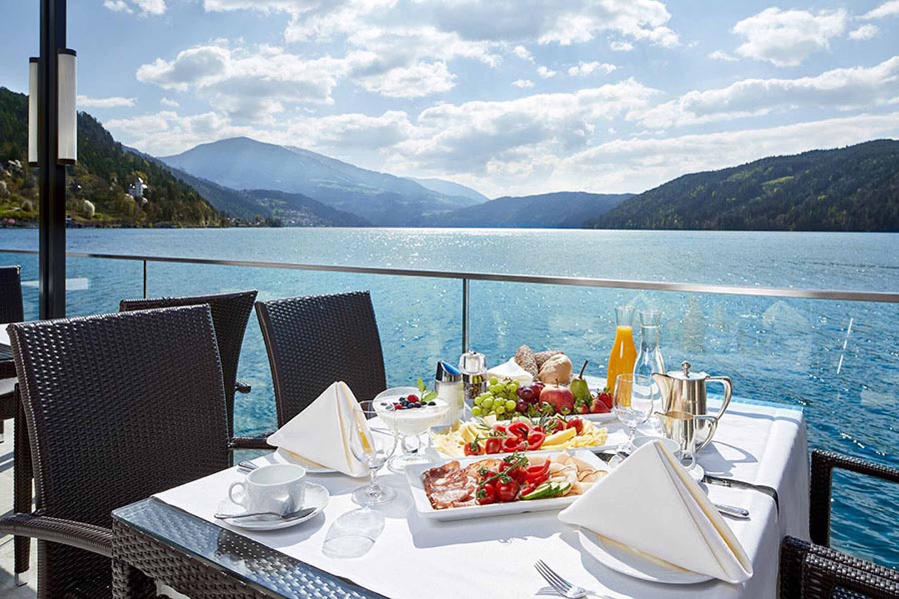 Una mesa lista para el desayuno con vistas a un lago, sillas de mimbre y montañas al fondo.