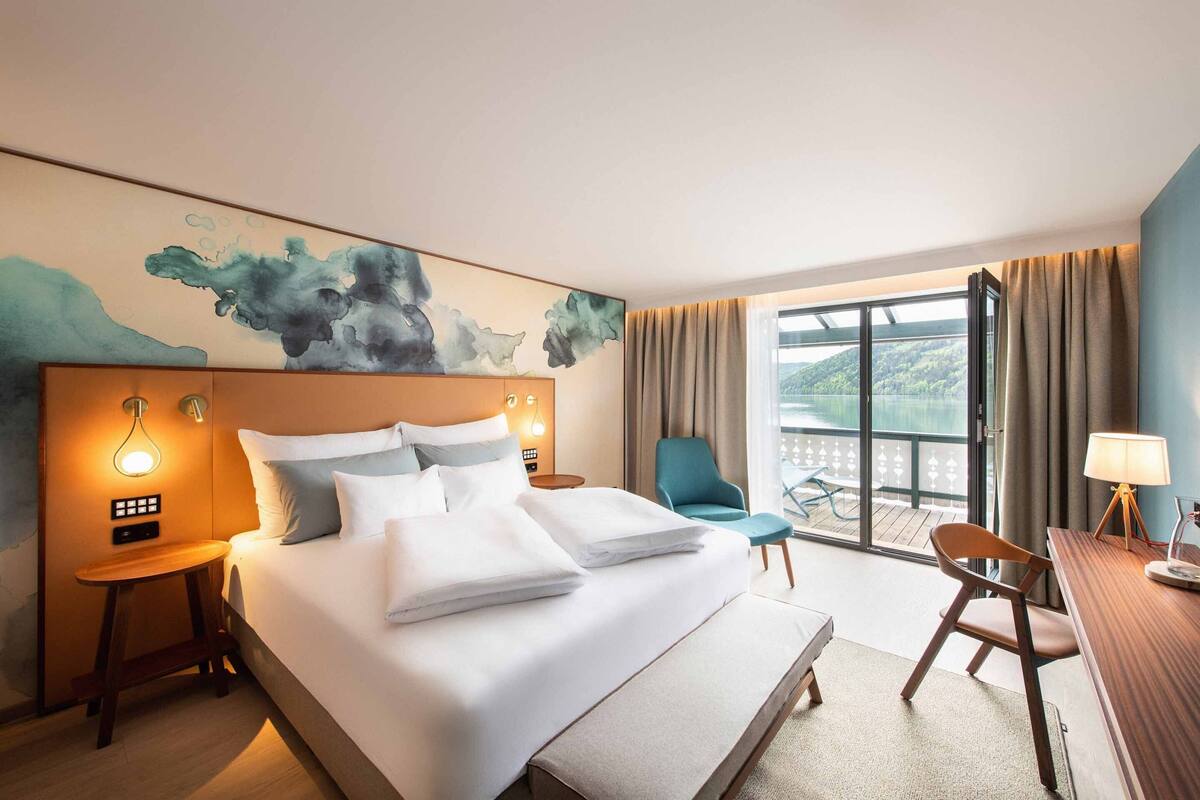 A hotel room with a large bed, a desk, a chair, a view of the outdoors, and a painting on the wall.