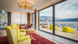 A modern hotel room with a large window overlooking a body of water and mountains. There is a yellow armchair, a red sofa, and a small round table with a vase. - Seeglück Hotel Forelle Superior (Millstatt)