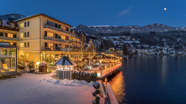 A hotel with a balcony overlooking a lake, snow-covered surroundings, and a mountain in the background. - Seeglück Hotel Forelle Superior (Millstatt)