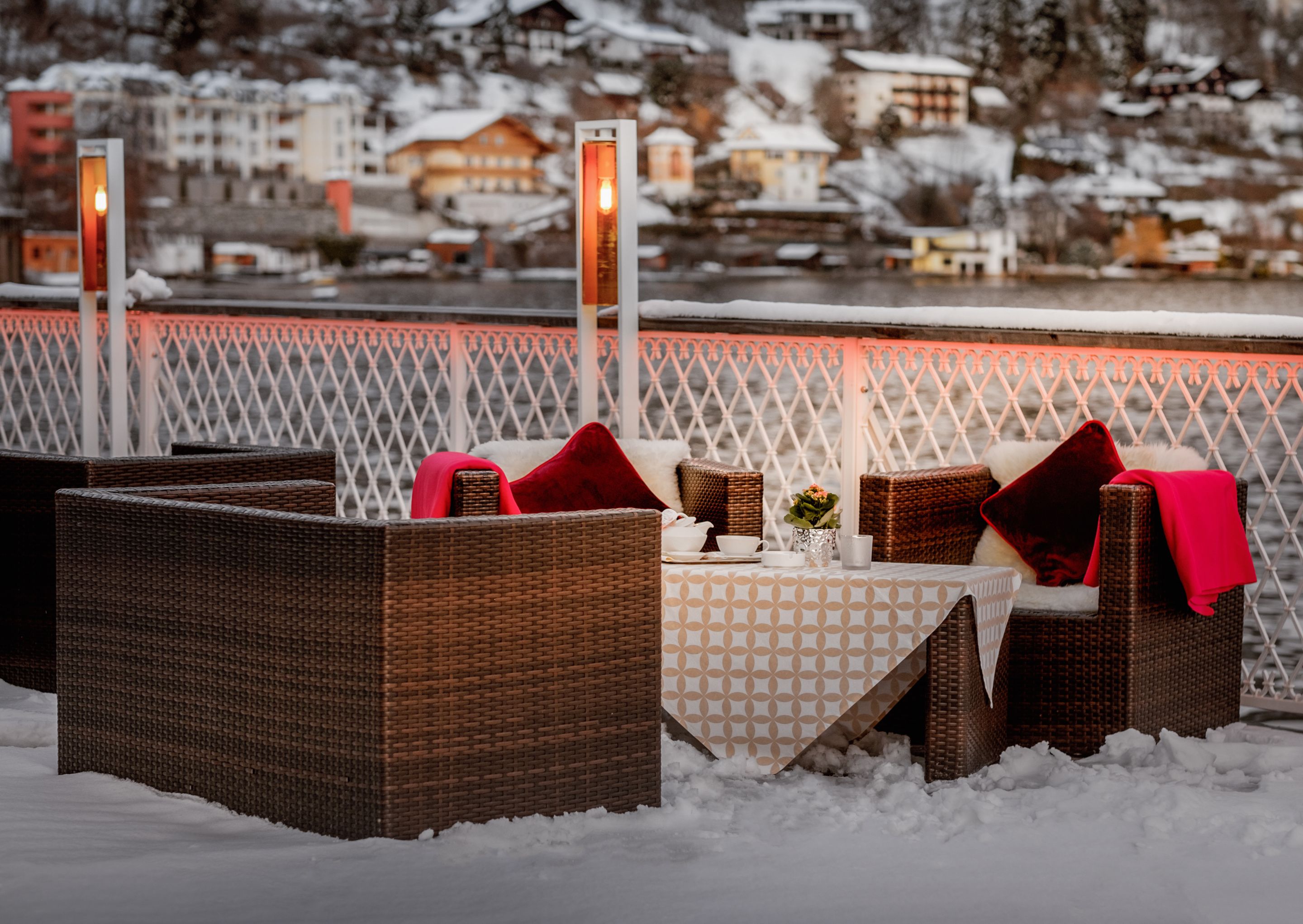 A snow-covered outdoor seating area with wicker furniture and a table set for dining, overlooking a snowy landscape with buildings in the background.
