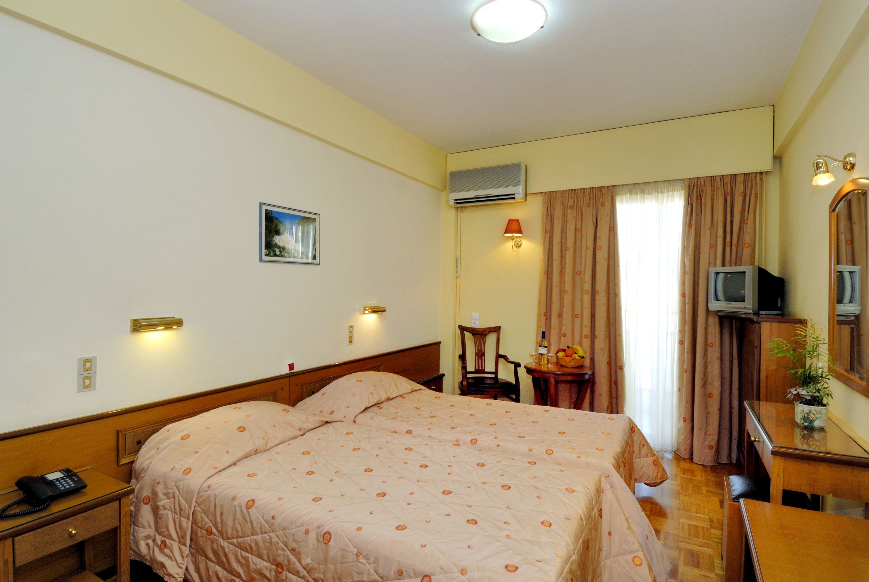 A hotel room with a double bed, a desk with a telephone, a chair, a small table with a plant, and a television on a wooden cabinet.