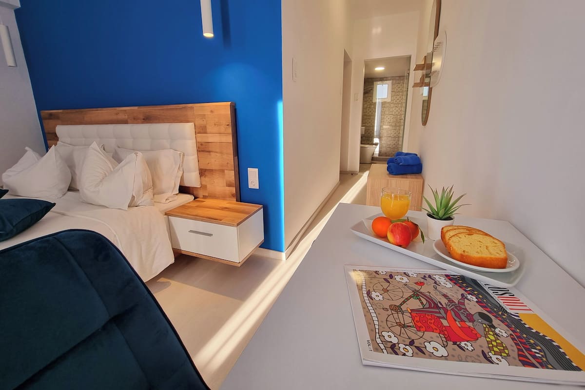 Un dormitorio moderno con una pared azul de acento, un cabecero de madera, una mesita de noche con lámpara y una bandeja de desayuno con fruta y pan.